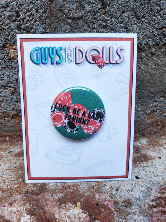 A GUYS AND DOLLS "Luck Be A Lady" Metal Pinback Button by The Lobby Boutique with the words 'guys and dolls' on it, featuring chips and dice.